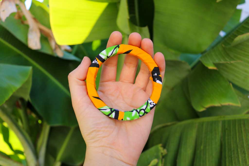 Uma mão a segurar a pulseira bangle azul com padrão africano sobre base rígida de madeira reaproveitada