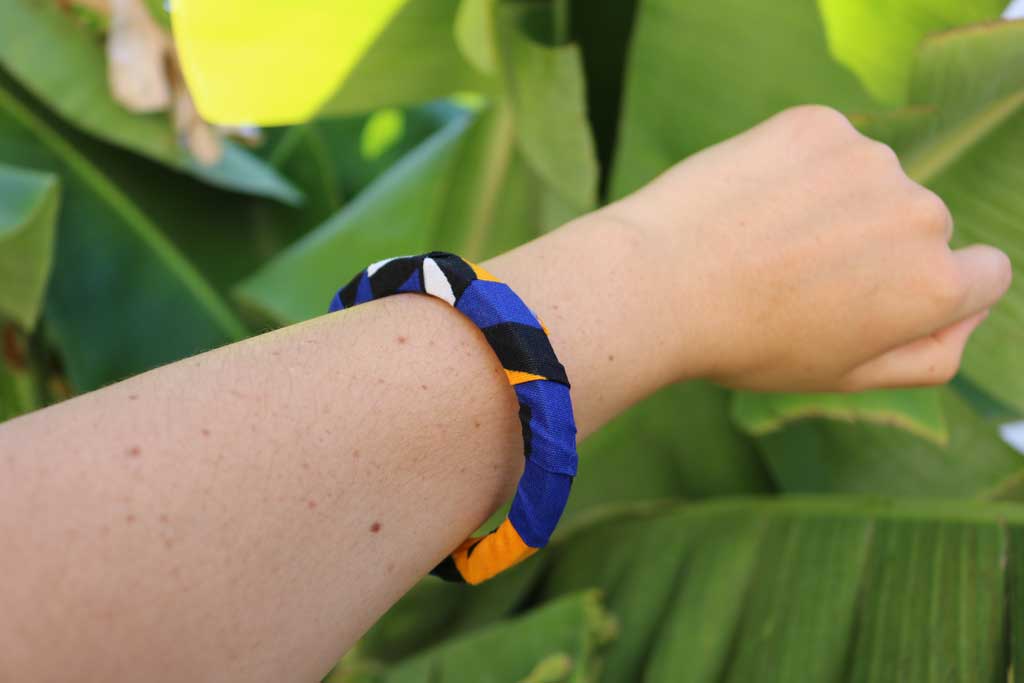 Uma mão a segurar a pulseira samakaka com padrão africano sobre base rígida de madeira reaproveitada