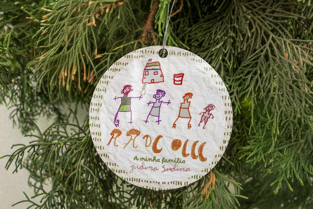 Enfeite de árvore de Natal feito em papel com desenho infantil de várias pessoas diante de uma casa